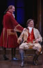 Archer in long red coat with gold embroidery and riding boots, his hair in a pony tail, leans on  the chair in which sits Aimwell, in tan coat and britches, red vest, and fluffy white shirt, black riding boots with brown tops, and long hair. Next to him is a table with pewter ewer and tankards